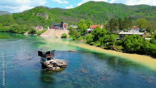 Aerial view of Bajina Bašta and the iconic Kućica na Drini (Drina River House), peacefully perched on a rock in the middle of the emerald Drina River, surrounded by lush nature and mountain landscapes