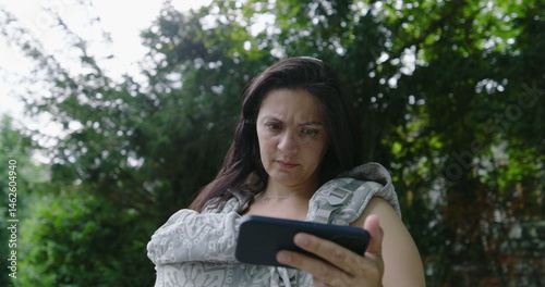 Mother with a preoccupied expression, holding her baby in a front carrier while intently focusing on her smartphone in a park, highlighting the blend of modern concerns and parenting