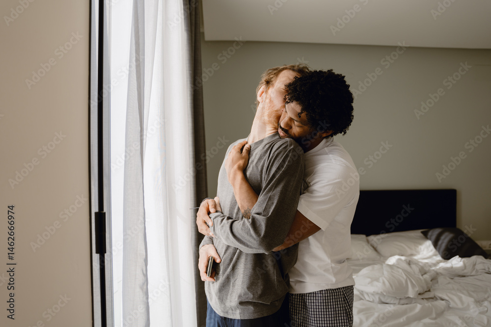 Fototapeta premium A man stands and hugs a man standing in front of him from behind, in a bedroom