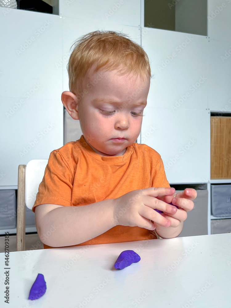 A red-haired boy in an orange T-shirt sculpts from colored plasticine at a table in a room. Development of fine motor skills according to the Montessori system