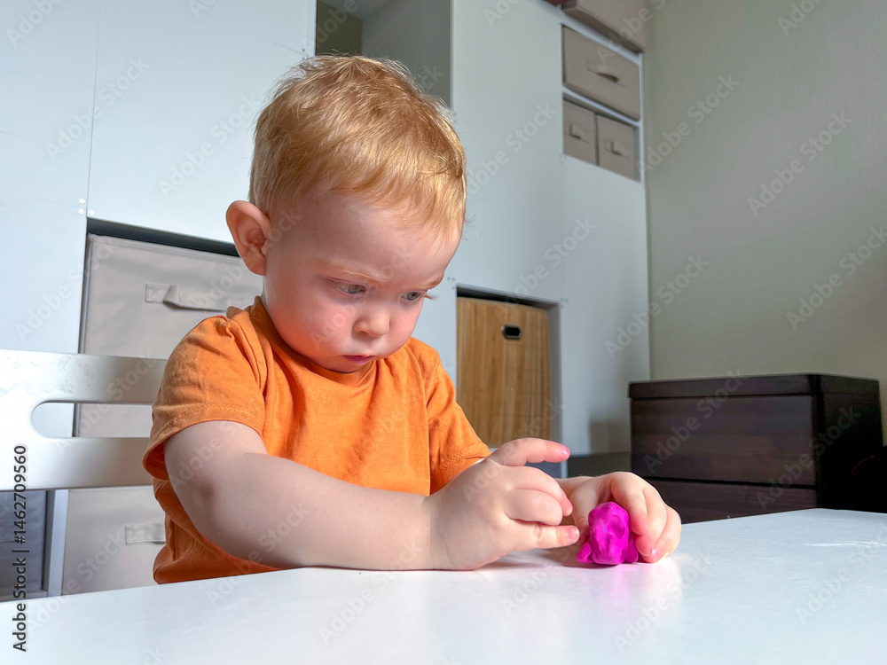 A little red-haired boy is sitting and sculpting from colored plasticine at a table in a room. Developing fine motor skills at the table