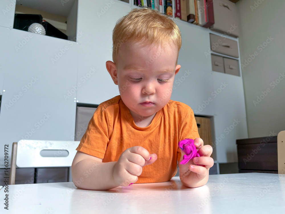 A red-haired boy in an orange T-shirt sculpts purple plasticine at a table. Early development of a child at home