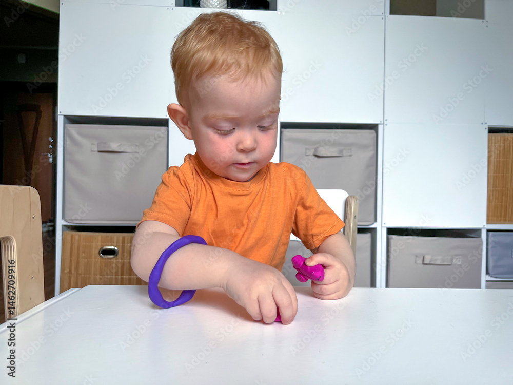 A little red-haired boy with a ring made of plasticine on his hand is playing at a table with plasticine. Early development of fine motor skills, creativity