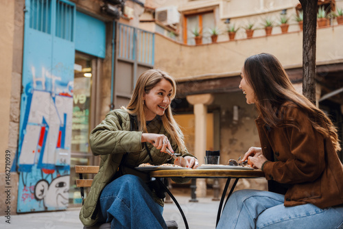 Two female friends sitting at a table and laughing while holding pizza knives, on the terrace of a restaurant