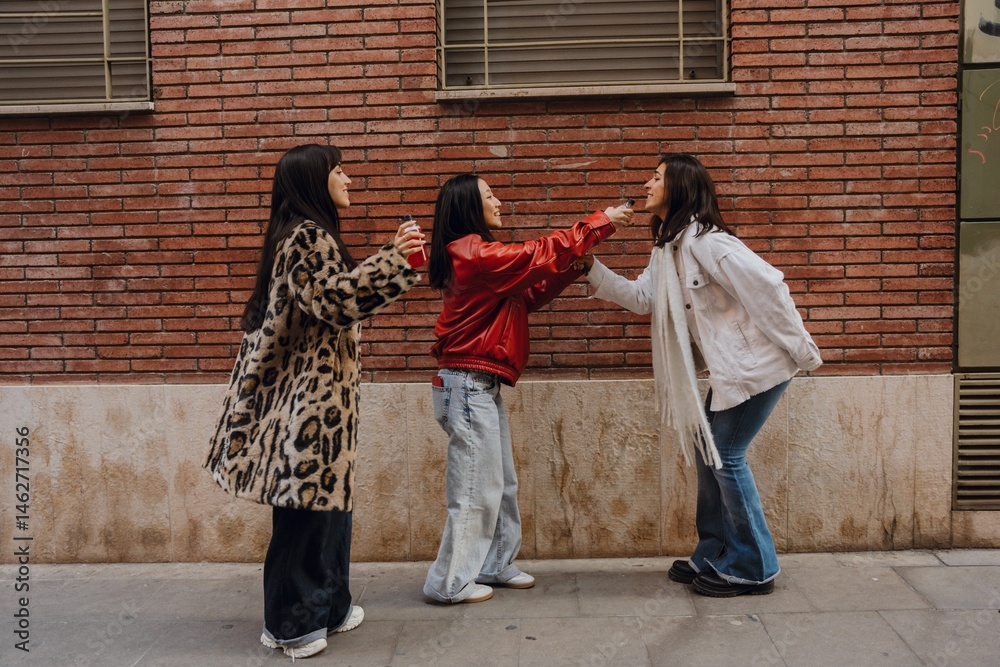 Naklejka premium A woman holds a bottle near the face of her female friend who is standing across from her while a female friend stands next to them, on the street