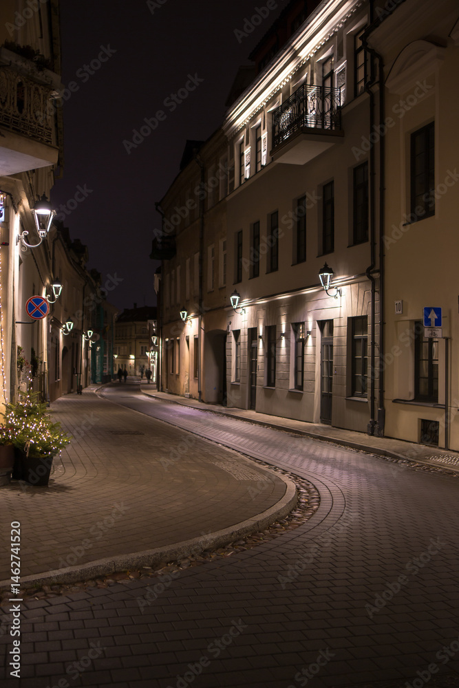 Obraz premium A vertical photo of a winding cobbled street in Vilnius Old Town at night. On both sides of the street are old buildings with illuminated windows and decorative lanterns on the walls. 