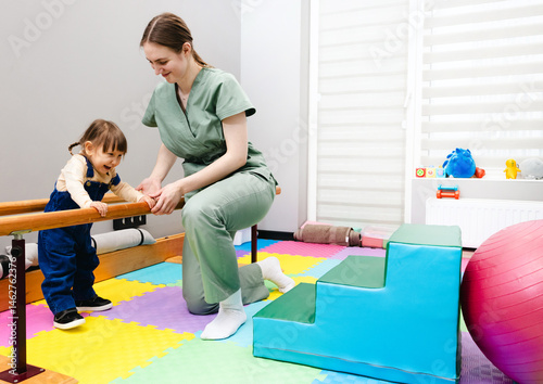 Young cute girl with cerebral palsy practices standing and balancing using handrails with assistance from physical therapist in a colorful therapy room