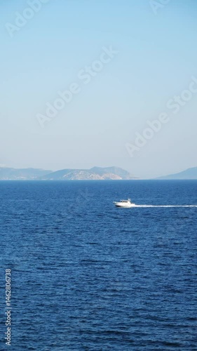 A white boat sailing on the sea, leaving a trail behind, with distant mountains in a blue haze. Captured in Athens, showcasing serene waters and a peaceful landscape.