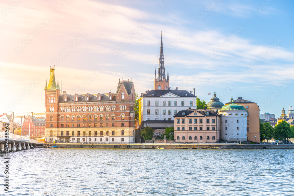 Naklejka premium Riddarholmen Island showcases beautiful architecture along the water's edge in Stockholm. The sun casts gentle light, highlighting historic buildings and a serene atmosphere.