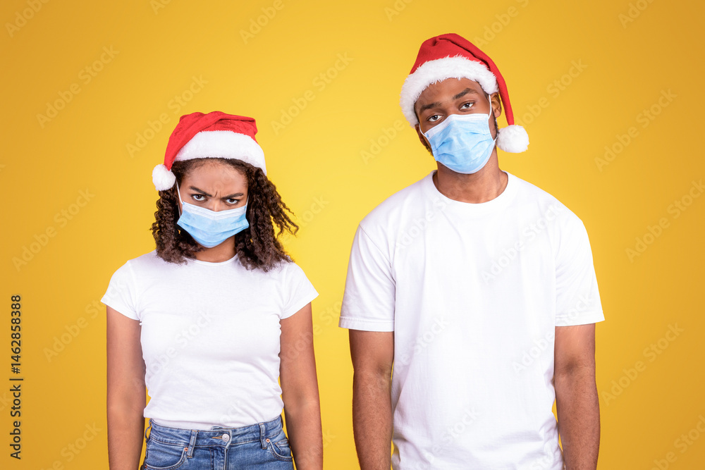 Fototapeta premium Coronavirus Christmas. Frustrated African American Couple Wearing Santa Hats And Protective Medical Masks Looking At Camera Standing On Yellow Studio Background. Xmas Party Cancelled Due To Covid-19