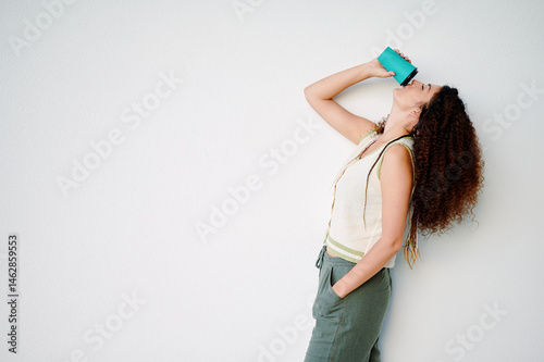 woman with curly hair leans against wall while drinking from reusable cup, symbolizing sustainability and modern lifestyle. casual outfit and relaxed pose convey everyday comfort. photo