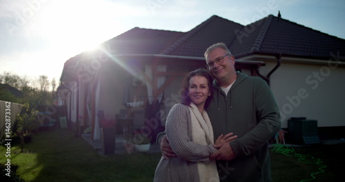 Couple embracing in backyard, standing near residential house, warm and affectionate moment in suburban home setting with patio, lawn, and greenery