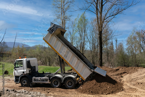 White dump truck unloading soil at a construction site. Heavy machinery performing earth works, backfilling for a landscape architecture project. Industrial earthmoving operation.