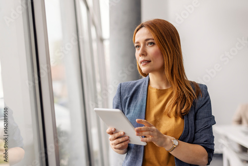 Photos Female business expert with tablet in hands looking through the window while standing at corporate office