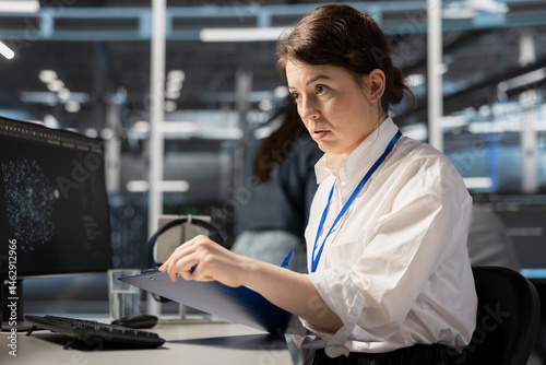 Data center admin monitoring system performance using PC, looking over maintenance paperwork. Server farm worker reading documentation files to implement optimizations