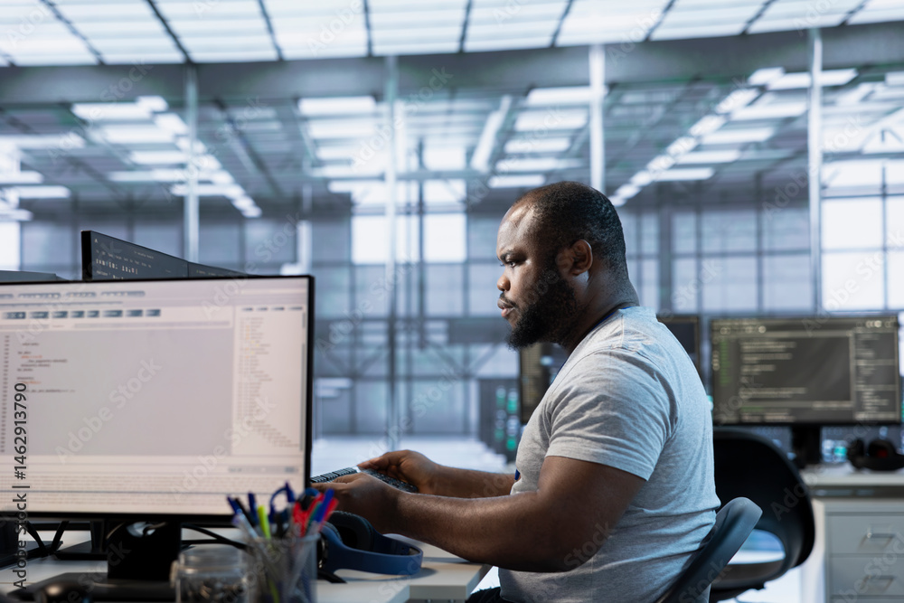 Technician at desk writing code and doing checkup on server room providing computing resources for different workloads. African american man investigating data center mainframes using computer