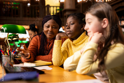 Obraz na płótnie African american professor mentoring her teen girls students with an assessment, teaching academic concepts and enhancing their knowledge through practical exercises. Private lessons at the library. w salonie