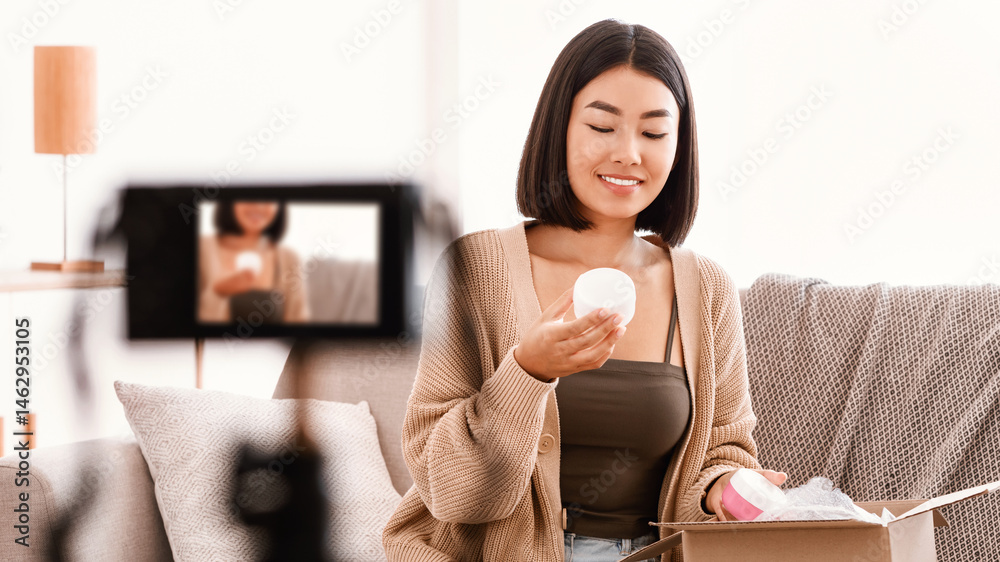 © Prostock-studio - Advertising Content. Asian young female influencer vlogger unboxing delivery with cosmetics product, holding jar with moisturizer cream, filming video for her beauty blog on camera at home