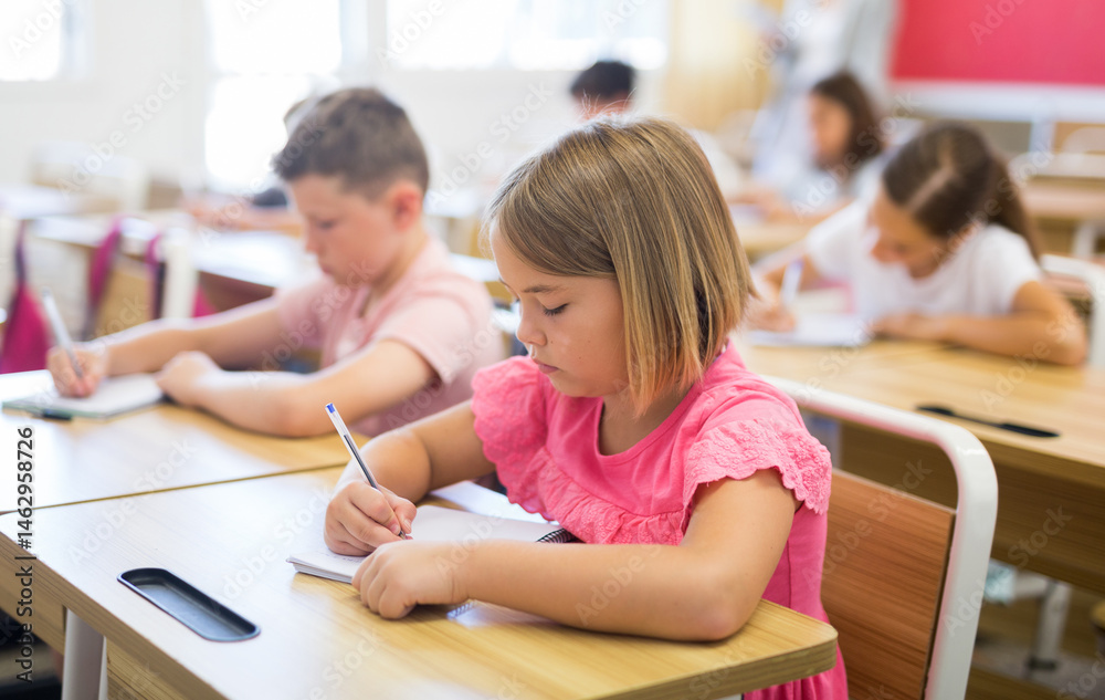 Portrait of focused cute small schoolgirl writing exercises in workbook during lesson in classroom .. Portrait of focused cute small schoolgirl writing exercises in workbook during lesson in classroom ..