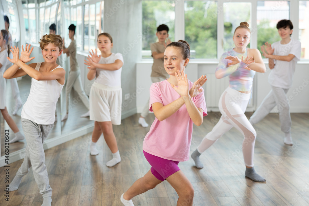 Fototapeta premium Female teen performs choreographic exercises and teaches energetic mobile social house dance together with friends. Young girls and guys in repeat movements, train in spacious studio