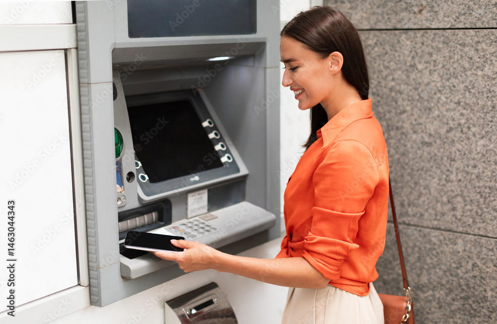 Young lady customer using smartphone to gain access to ATM, withdrawing money from account standing near bank machine on a street outside, side view shot. Financial safety and transactions Young lady customer using smartphone to gain access to ATM, withdrawing money from account standing near bank machine on a street outside, side view shot. Financial safety and transactions
