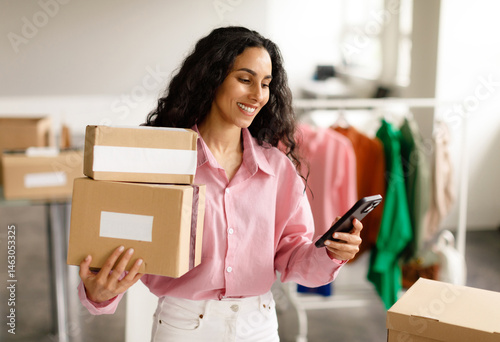 Clothing Shop Entrepreneurship. Happy Lady Using Smartphone Holding Packed Cardboard Boxes With Clothes And Outfits, Sending Message To Buyer Standing In Store. Small Retail Business, Ecommerce