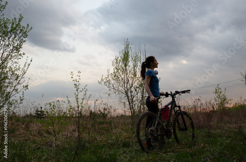 Young sporty woman riding a bicycle on the spring field with green grass on sunset sky landscape background. Happy activity sport trip vacations