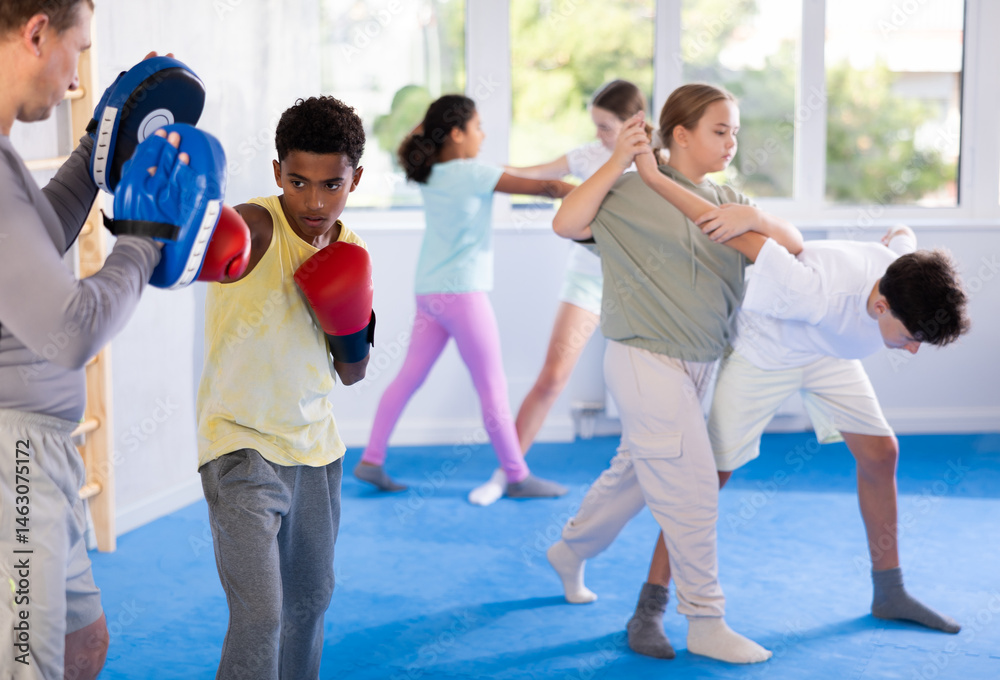 Obraz premium Motivated focused african american boy wearing boxing gloves working out with coach during group self-defense course, practicing punches on mitts..