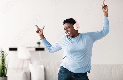 Music Lover Concept. Overjoyed ecstatic plus-size african american lady wearing pink wireless headphones listening to favourite song, dancing at home with closed eyes. Female taking break, copy space photo