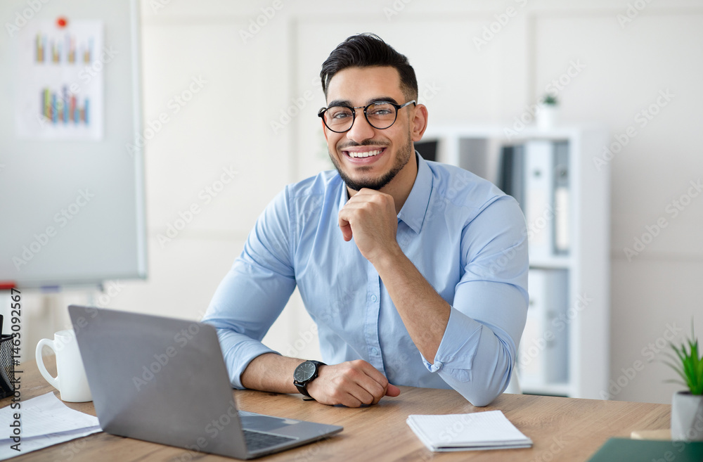 Portrait of happy Arab businessman using laptop computer, making video call, chatting online with colleague at office. Young male entrepreneur having virtual meeting or business webinar