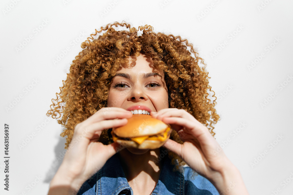 Young woman with curly hair joyfully holding a burger, showcasing healthy eating and enjoyment of fast food Bright background emphasizes the playful mood