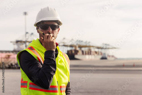 A port worker wearing a safety vest, hard hat, and sunglasses uses a walkie-talkie while overseeing cargo operations, with large container cranes and ships visible in the background at the dock.