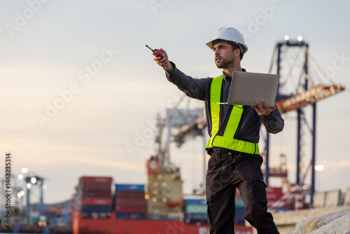 A male port engineer wearing a safety vest and helmet holds a laptop and talks on a radio at a busy shipping dock, overseeing cargo operations with cranes and containers in the background.