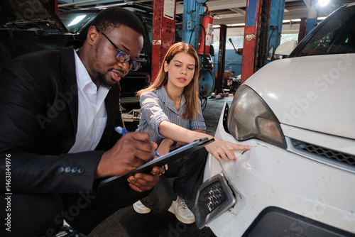 Warranty service business, Black male insurance agent inspector records crash car damage bumper, checks list and explains claim comprehensive of wreck fix to White female customer at auto garage.