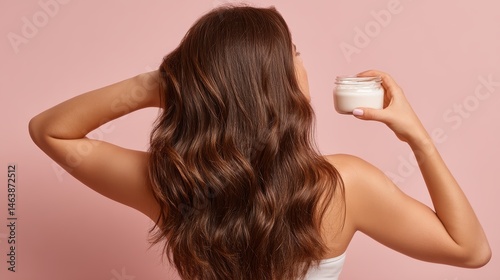 Woman applying hair mask to her long hair, back view, with a jar in hand and a light pink background