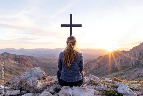 Woman meditates before a cross at sunset atop a mountain, overlooking a vast desert landscape
