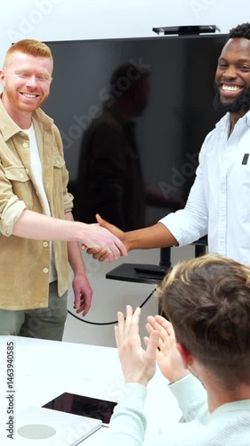 Professional businesspeople celebrating successful partnership through firm handshake, colleagues clapping in bright, modern office setting, signaling teamwork achievement