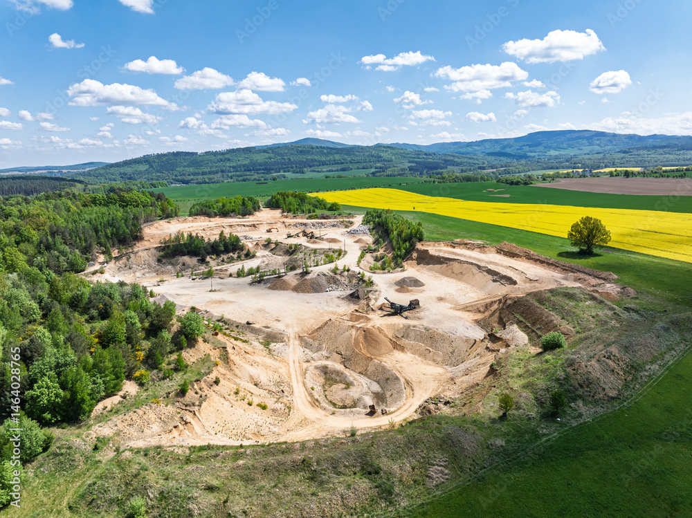 Aerial drone view of sand or gravel quarry surrounded by forest and farmland with distant hills. Aerial Drone view shows an inactive or abandoned quarry embedded in a green landscape.