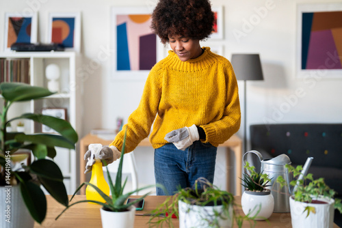 A woman in a yellow sweater tends to her indoor plants. She is using a spray bottle to water them in a bright, modern living room.
