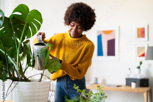 A woman in a yellow sweater waters her Monstera plant with a silver watering can. She is smiling and taking care of her indoor garden in her home.