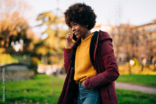 A smiling young woman with an afro is talking on her phone outdoors. She is wearing a yellow sweater and a maroon winter coat.