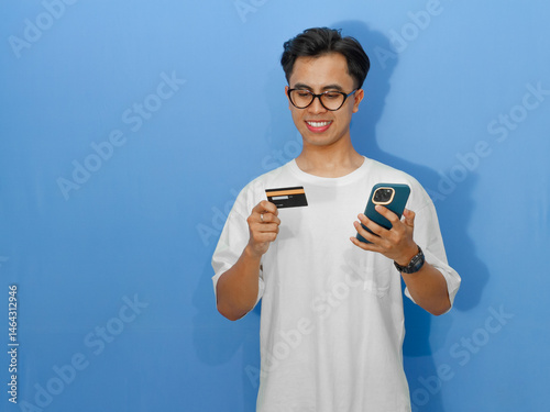 Happy young Asian man in a white t-shirt making an online payment with a mobile phone and credit card.