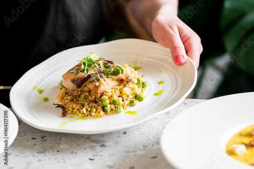 Wallpaper Mural A hand holding a white plate with a gourmet dish featuring a perfectly cooked fish fillet atop grains and green peas, garnished with microgreens and drizzled with a vibrant green sauce Torontodigital.ca