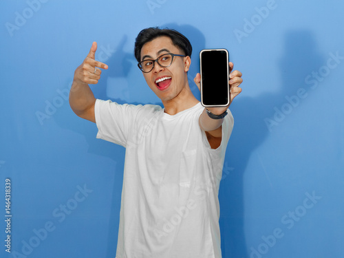 Excited young Asian man in a white t-shirt pointing at a blank mobile phone screen, perfect for promotion, advertising, or digital marketing content.