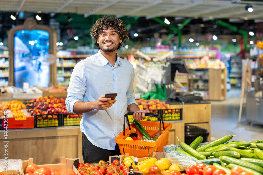 Obraz premium A smiling man at the grocery store holds a basket of produce and a smartphone, looking at the camera.