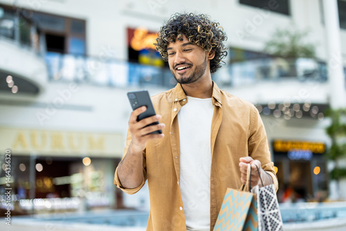 Canvastavla A smiling man looking at his phone while carrying shopping bags inside a modern shopping mall