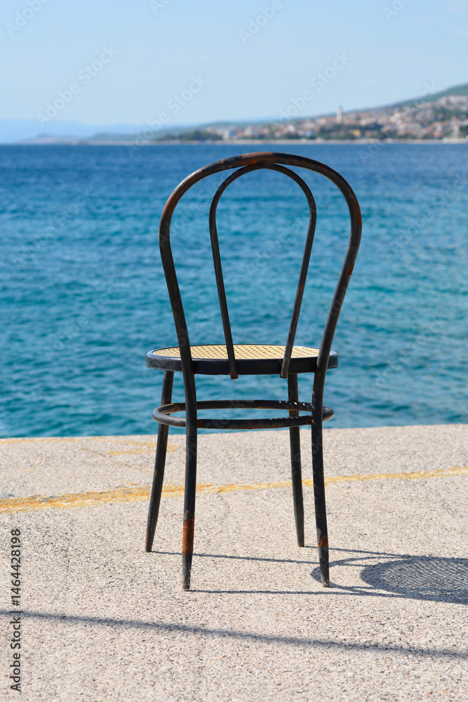 A classic dark wooden chair with a woven seat stands on the waterfront in Selce, with the distant cityscape of Crikvenica across the calm blue sea