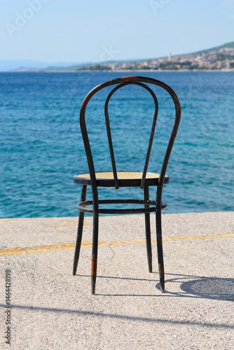 Obraz na płótnie A classic dark wooden chair with a woven seat stands on the waterfront in Selce, with the distant cityscape of Crikvenica across the calm blue sea w salonie