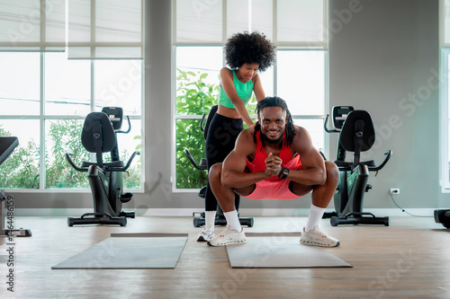 Black father doing squats on exercise mat while his daughter playfully pushes his shoulders, enjoying a fun workout session together, father daughter activity, healthy family