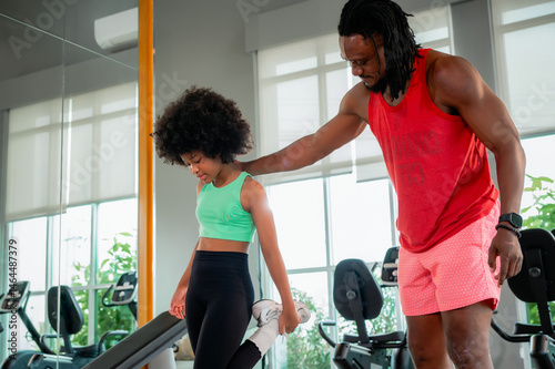 Black father as a fitness trainer helps his daughter stretch and balance in a modern gym filled with exercise equipment.